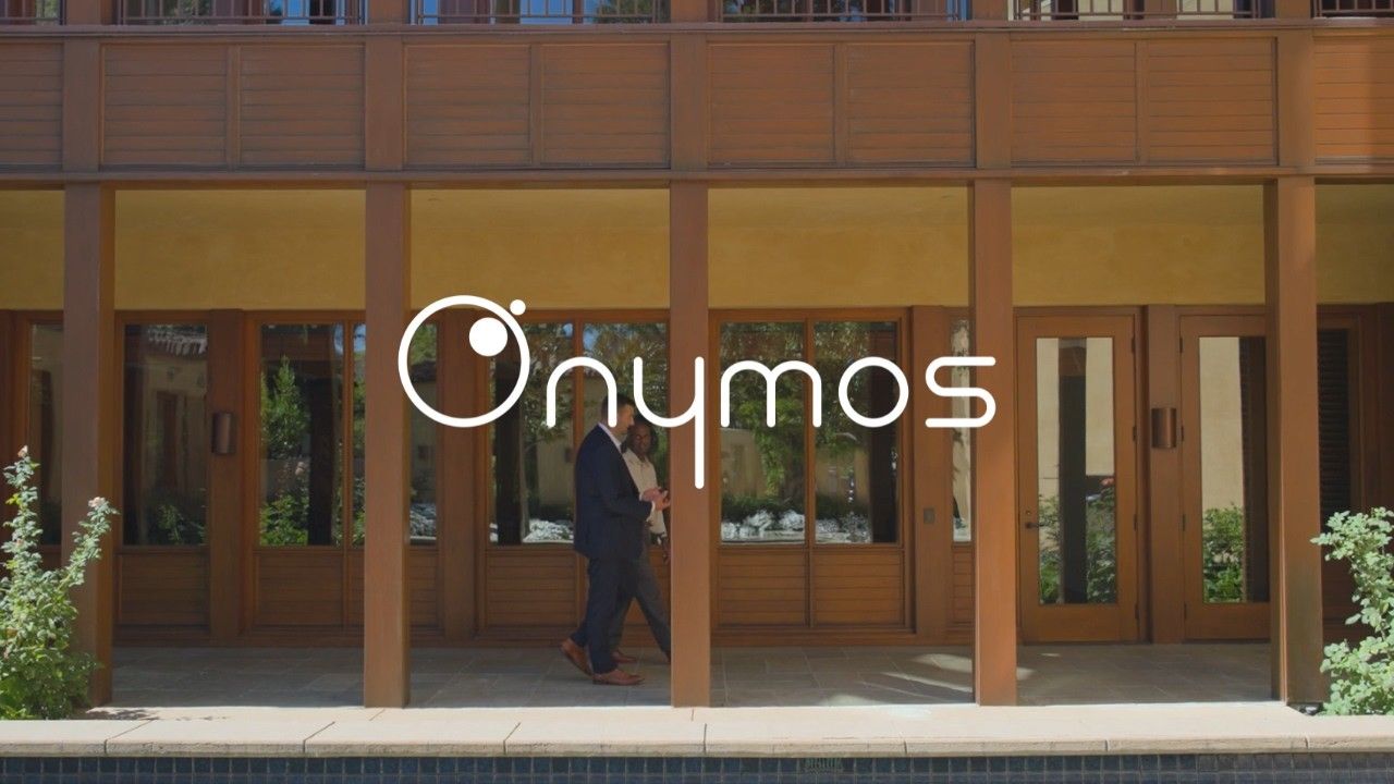 Onymos logo over two people walking past the wood-framed windows of an office courtyard